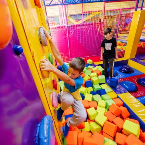 Children are playing in the indoor playground with a climbing structure, soft mats, and blocks.